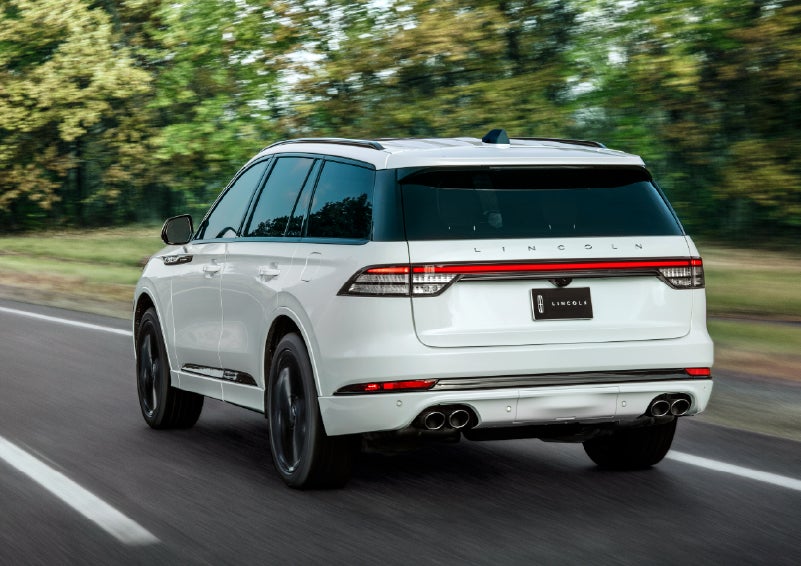 A white 2026 Lincoln Aviator® SUV is being driven away down a road lined with colorful trees. | Deacon Jones Lincoln in Goldsboro NC