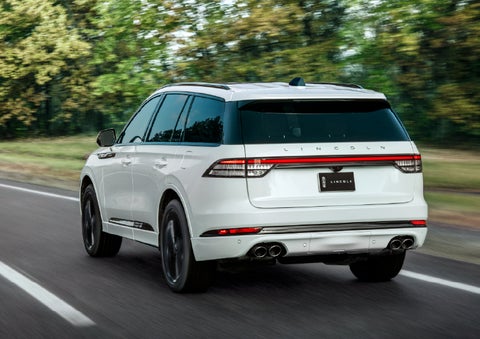 A white 2026 Lincoln Aviator® SUV is being driven away down a road lined with colorful trees. | Deacon Jones Lincoln in Goldsboro NC